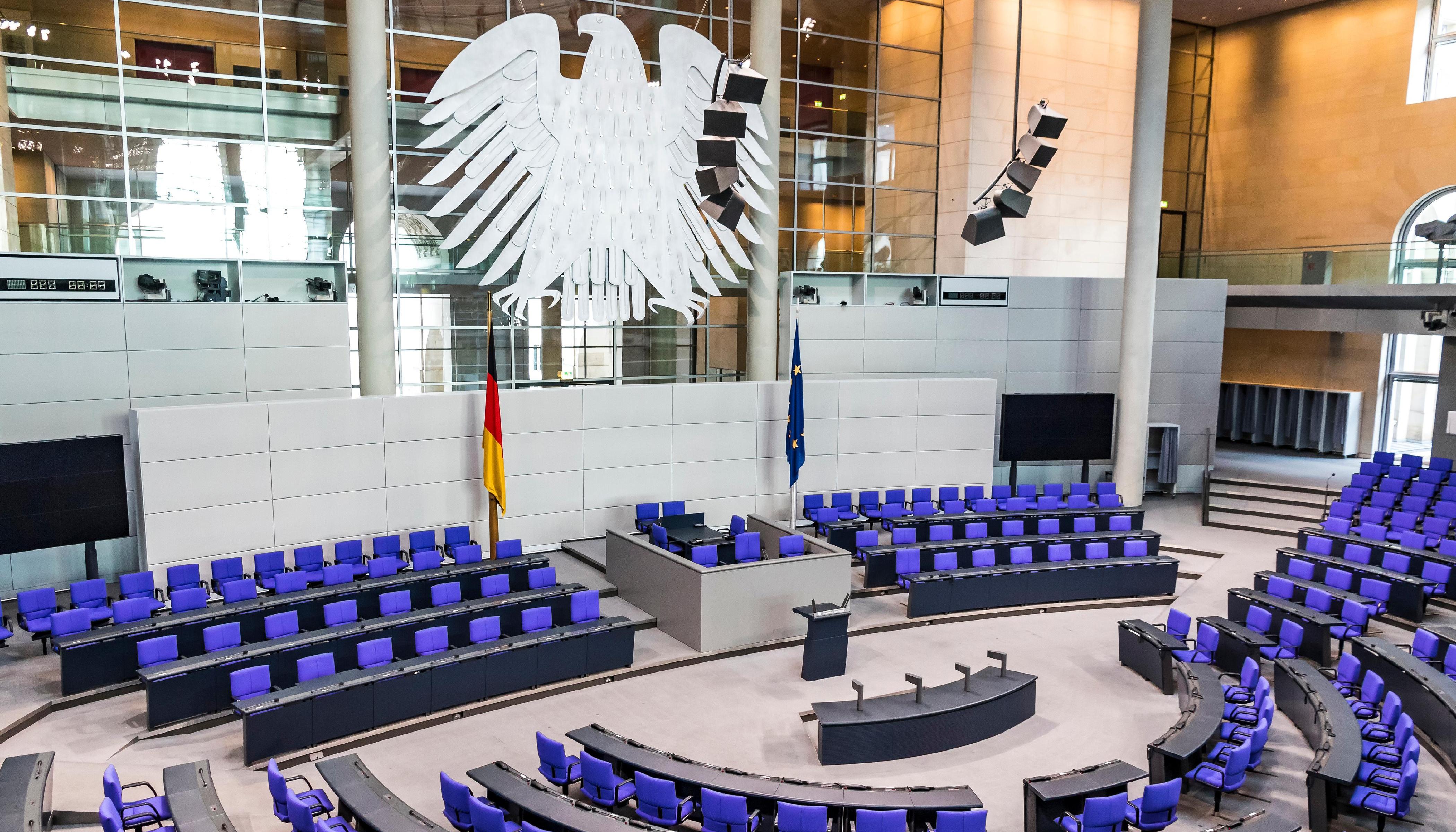 Eine Fotografie des leeren Plenarsaals des Deutschen Bundestages. Zu sehen ist die blaue Bestuhlung für die Fraktionen sowie die Plätze für die Regierung und die Paralmentspräsidenten. Darüber thront der Bundesadler. Quelle: stock.adobe.com/katatonia.