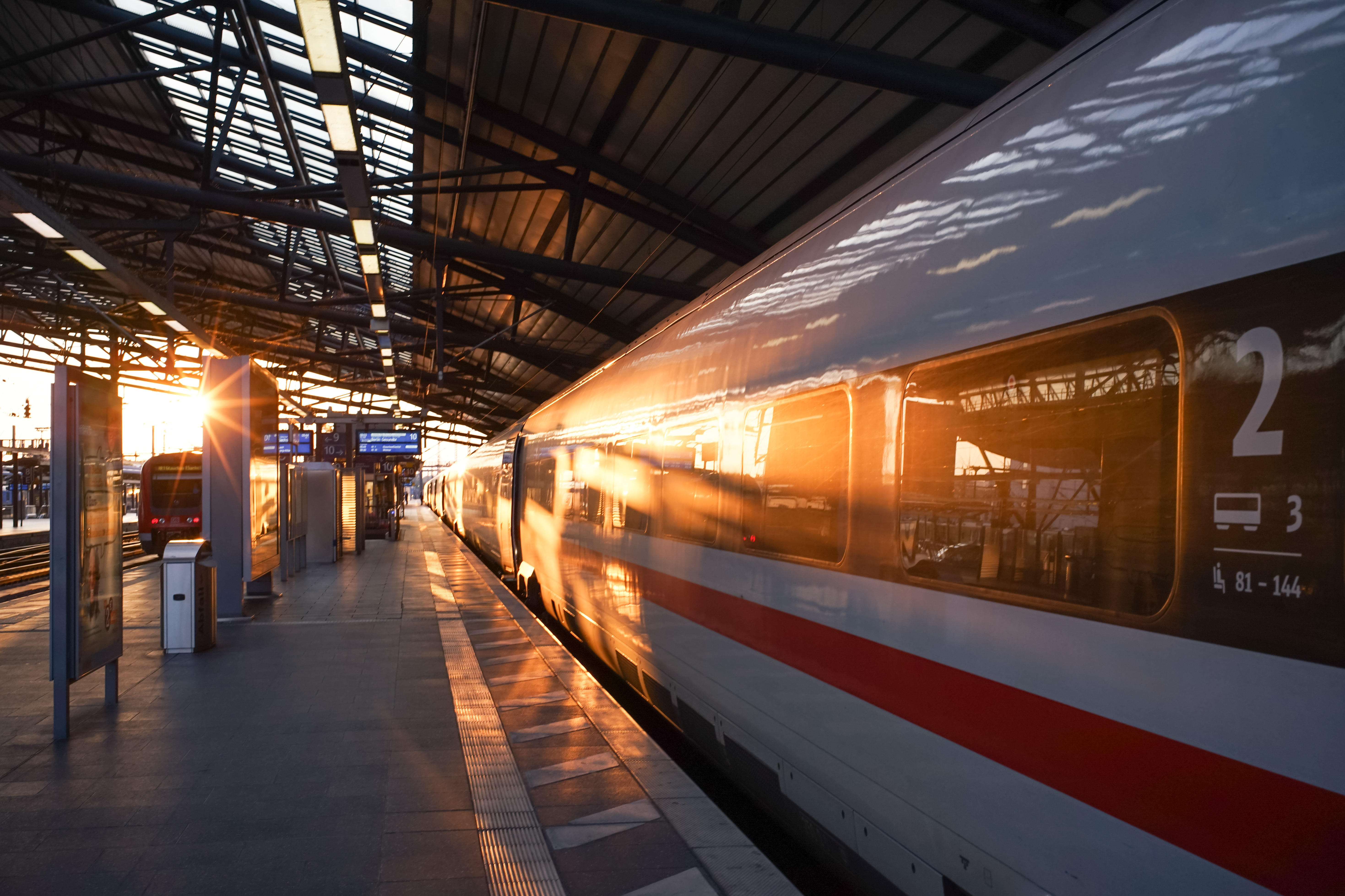 Ein ICE steht am menschenleeren, überdachten Bahnsteig. Auf dem gegenüberliegenden Gleis fährt ein Regionalzug in den Sonnenuntergang. Quelle: corofisch/stock.adobe.com.